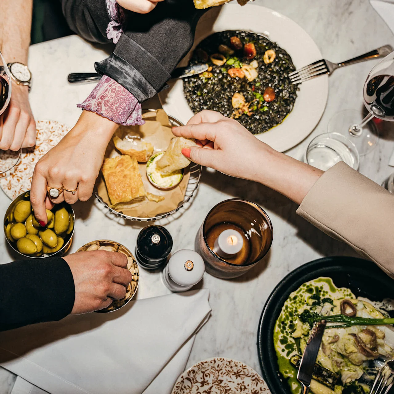 Several hands reach for food on a table. Olives, bread, and dishes including a green and black dish are present. Wine glasses and a lit candle add to the dining atmosphere.