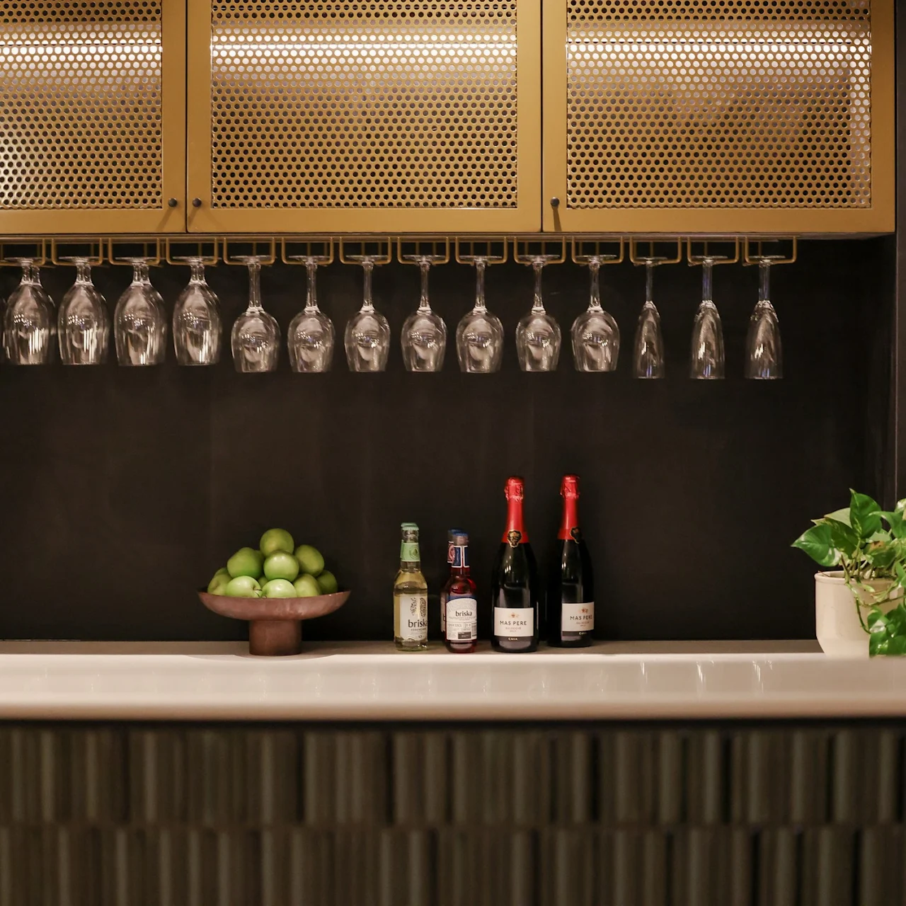 Hanging wine glasses under a shelf above a bar countertop lined with bottles, a bowl of green apples, and a potted plant in a stylish, modern setting.