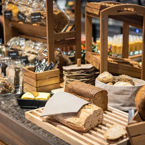 Brød ligger på en træbakke sammen med smør og krukker med pålæg. I en buffetopsætning er forskellige brød og tilbehør arrangeret med bestik i nærheden.