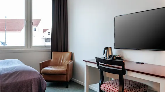 A well-lit hotel room features a brown leather chair beside a bed, against a window. A desk with a chair holds a kettle and a flat-screen TV is mounted on the wall.