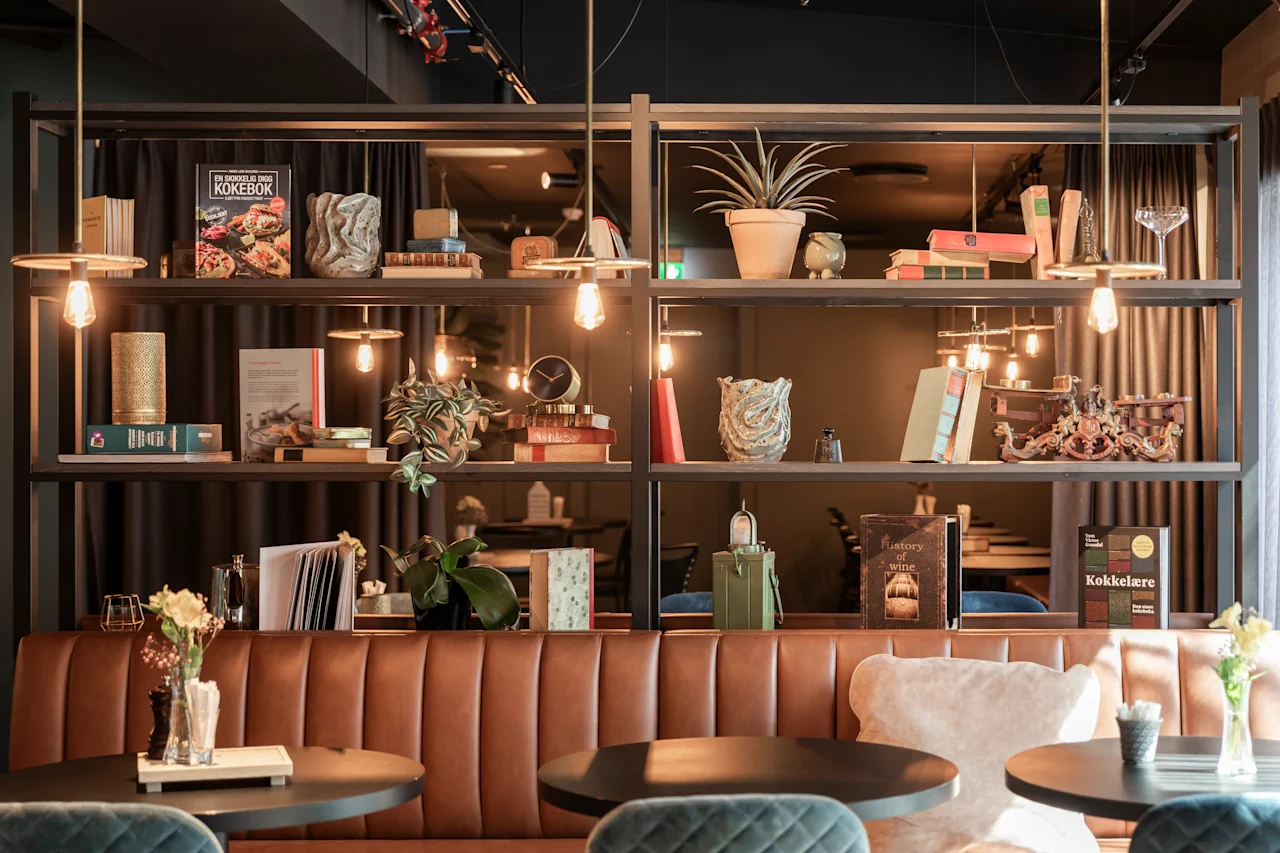 En mysig kafé med brun lädersoffa, runda bord och hyllor med växter och böcker. Varm belysning och små krukväxter skapar en inbjudande atmosfär. Synliga boktitlar: "KOKEBOK" och "Kokekatere".