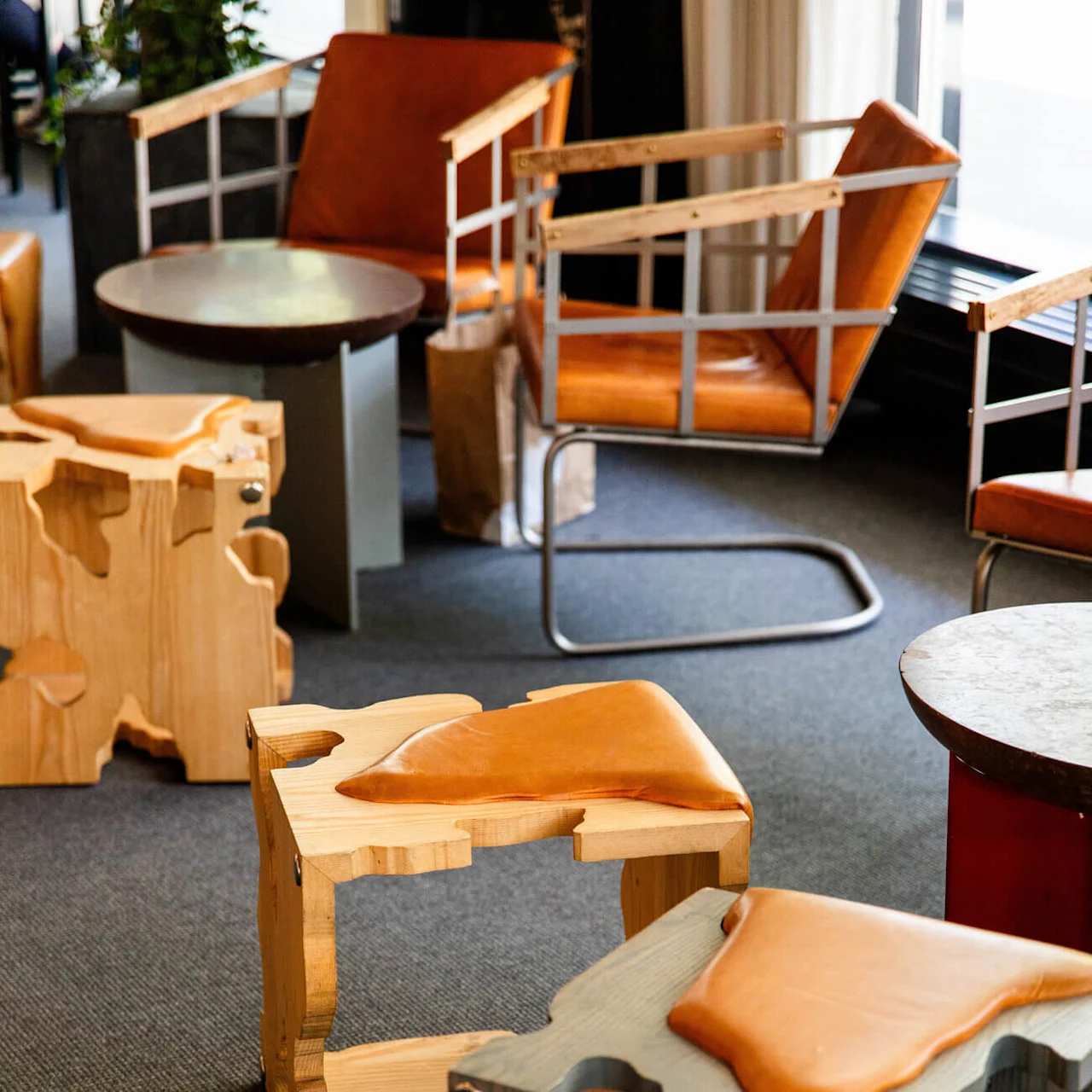 Wooden chairs with leather seats are arranged in a lounge area with dark carpet and metal-framed armchairs, creating a modern interior setting.