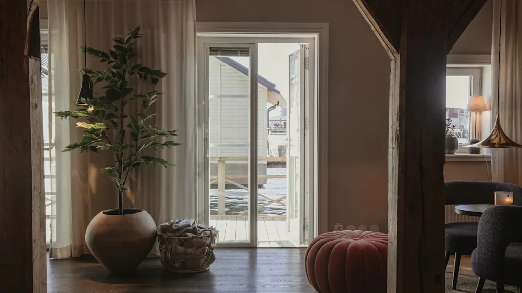 Et hyggeligt værelse med en stor potteplante og en rund puf, med udsigt gennem en åben dør til en vandudsigt med en bygning.