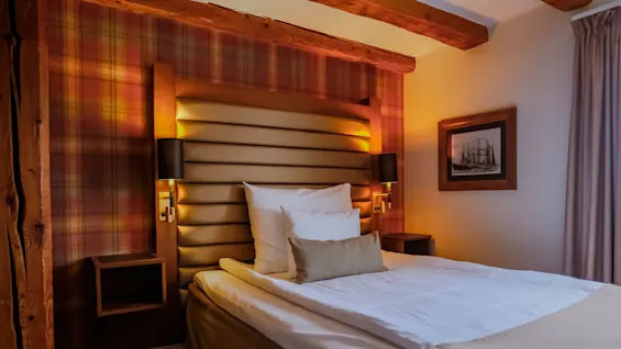 Et varmt og indbydende hotelværelse med en komfortabel seng, ternet tapet og synlige træbjælker, perfekt til et afslappende ophold.