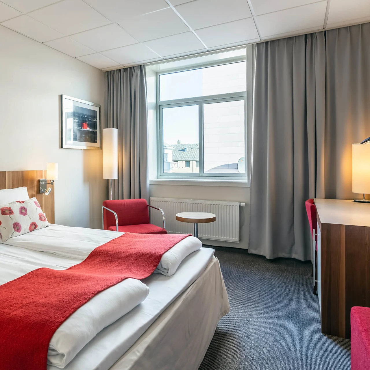 En pent hotelseng med røde detaljer står i et rom med et vindu, grå gardiner, en rød stol og et trebord under varm belysning.