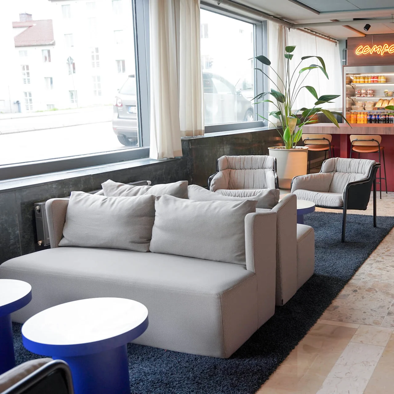 A modern lounge features gray sofas and chairs around small blue tables, with large windows and a neon sign reading "Comfort Deli". High stools line a bar counter, with shelves displaying snacks.