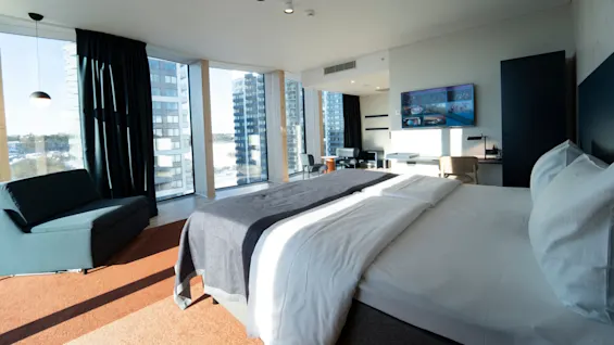 Bed with white linens in sunny hotel room, facing a wall-mounted TV. Large windows with curtains, couch, and chairs in modern decor. High-rise buildings visible outside.