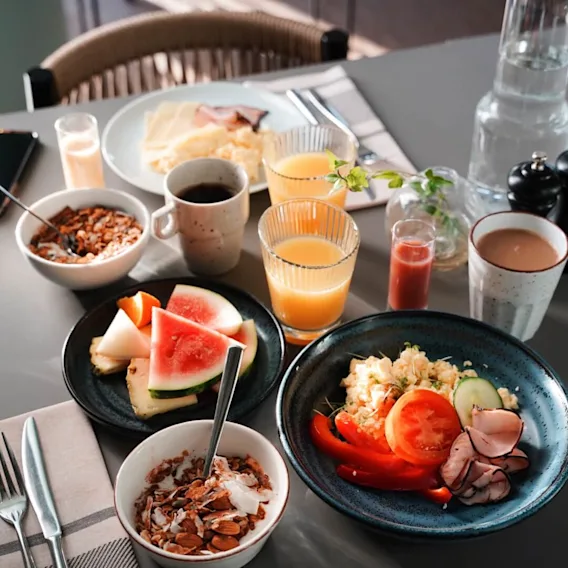 Frokostmat arrangert på et bord, inkludert eggerøre, frukt, yoghurt med nøtter, juice og kaffe; gjenstander er satt på en grå overflate med bestikk og en flaske med vann.