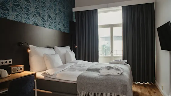 A neatly made bed with white linens and gray throw blanket is in a room. A wooden desk and chair are beside it. Dark curtains cover a window, with wallpaper behind the bed.