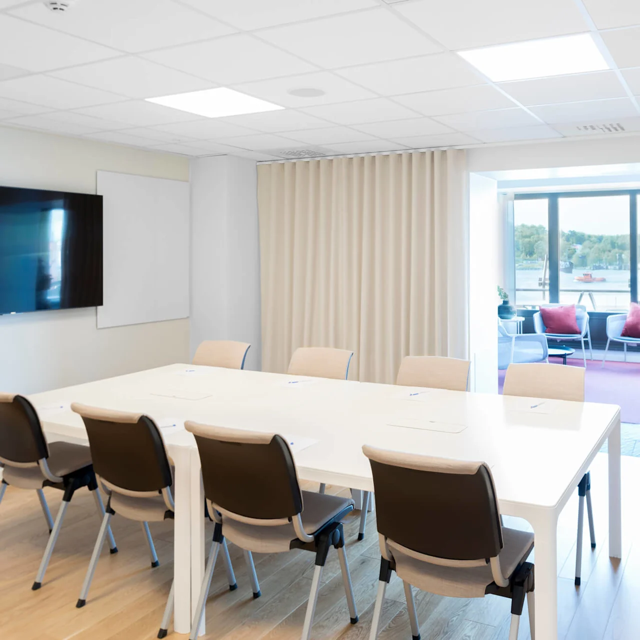 Conference room with a white table and six chairs, a large TV on the wall. Sunlit patio with chairs and a table is visible through glass doors.