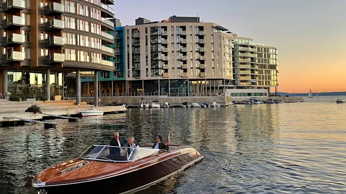 A wooden motorboat with three passengers navigates a calm harbor, surrounded by modern apartment buildings and a sunset-lit sky.