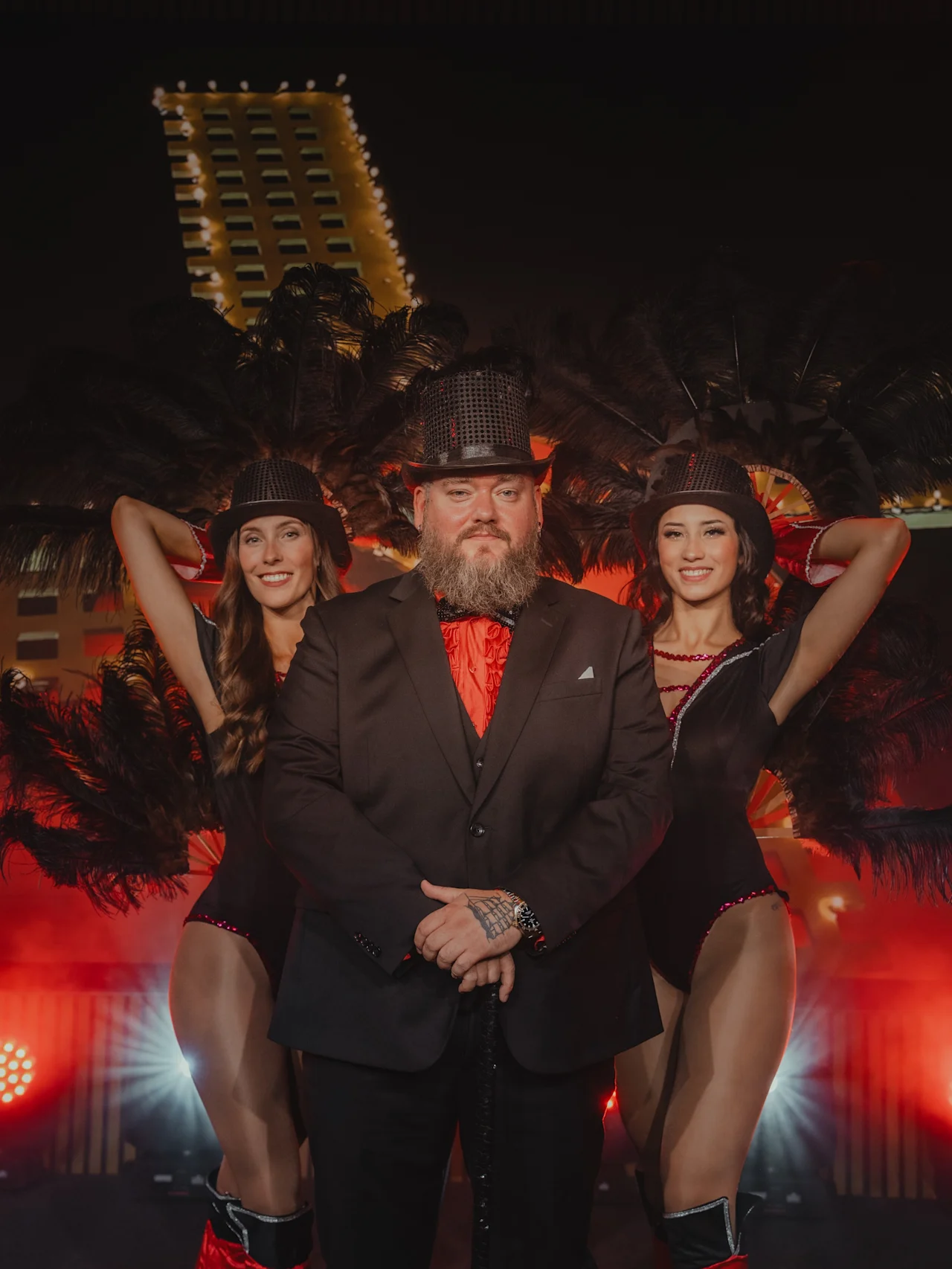 Mann i svart dress og hatt står med to kvinner i svarte drakter, røde støvler og hatter. De flankeres av fjærprydninger, med en opplyst bygning i bakgrunnen.