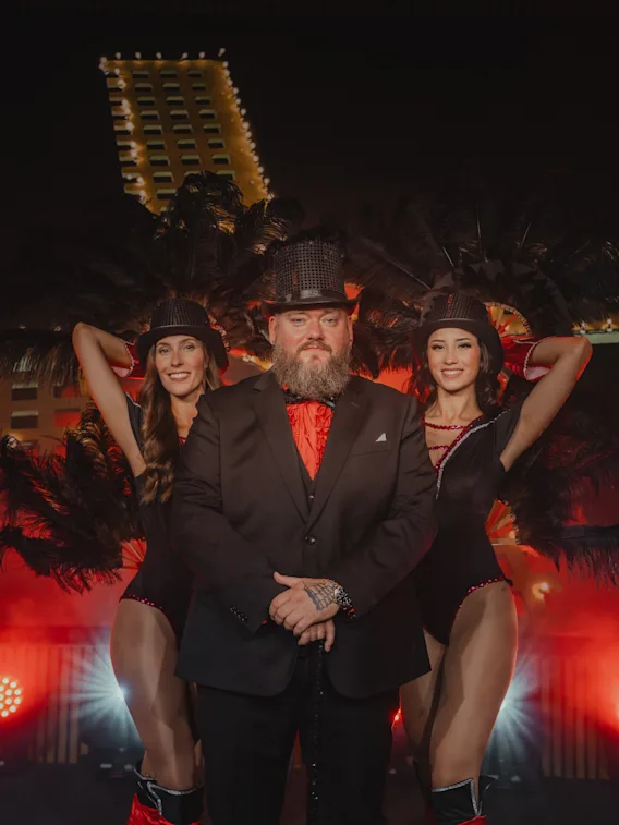 Man in a black suit and hat stands with two women in black costumes, red boots, and hats. They're flanked by feathered props, with a lit building visible in the night background.