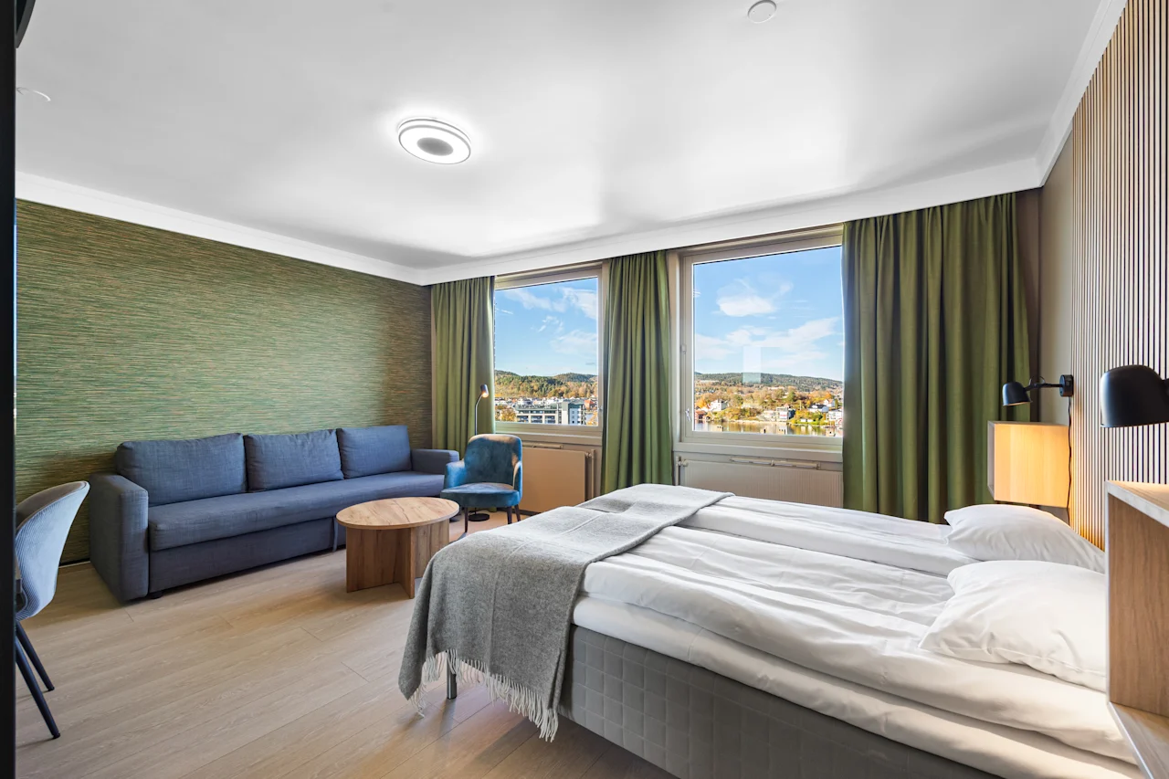 Lyst hotellrom med dobbeltseng, sofa og vinduer med utsikt.