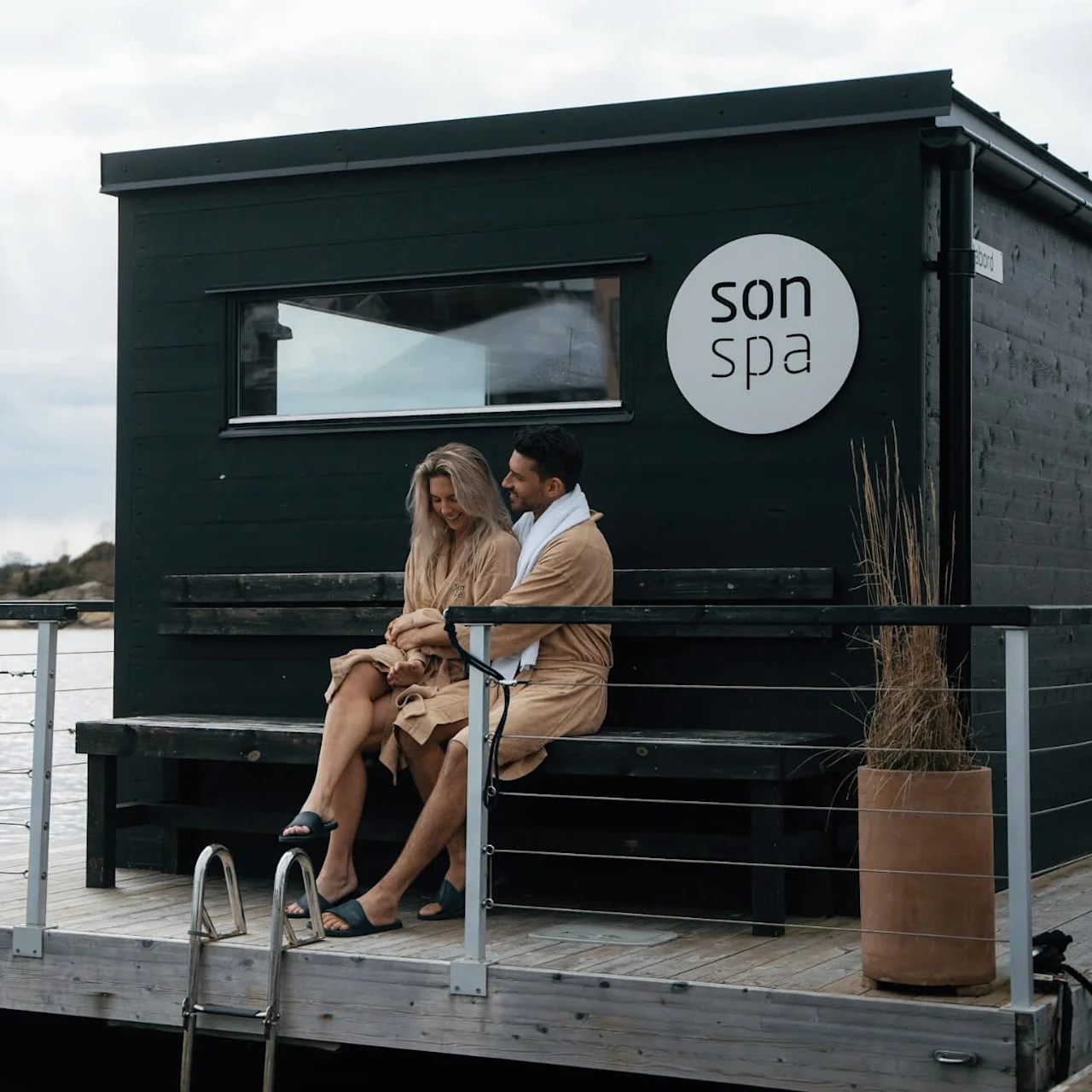 To personer i badekåper sitter på en benk ved en svart bygning merket "son spa," ved rolig vannkant.