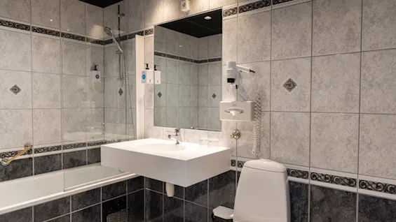 A bathroom with a white sink and mirror mounted on a tiled wall, a bathtub with a showerhead on the left, and a toilet on the right.