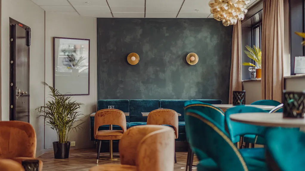 Velourlænestole omkring borde i en stilfuld lounge med dekorativ belysning og planter.