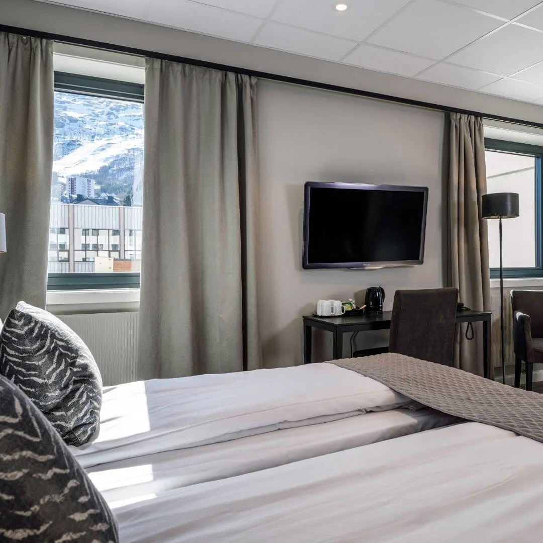 Et moderne hotelværelse har en stor seng med mønstrede puder, et vægmonteret tv, et lille bord, stole og en køjeseng. Vinduerne viser udsigt over snedækkede bjerge udenfor.