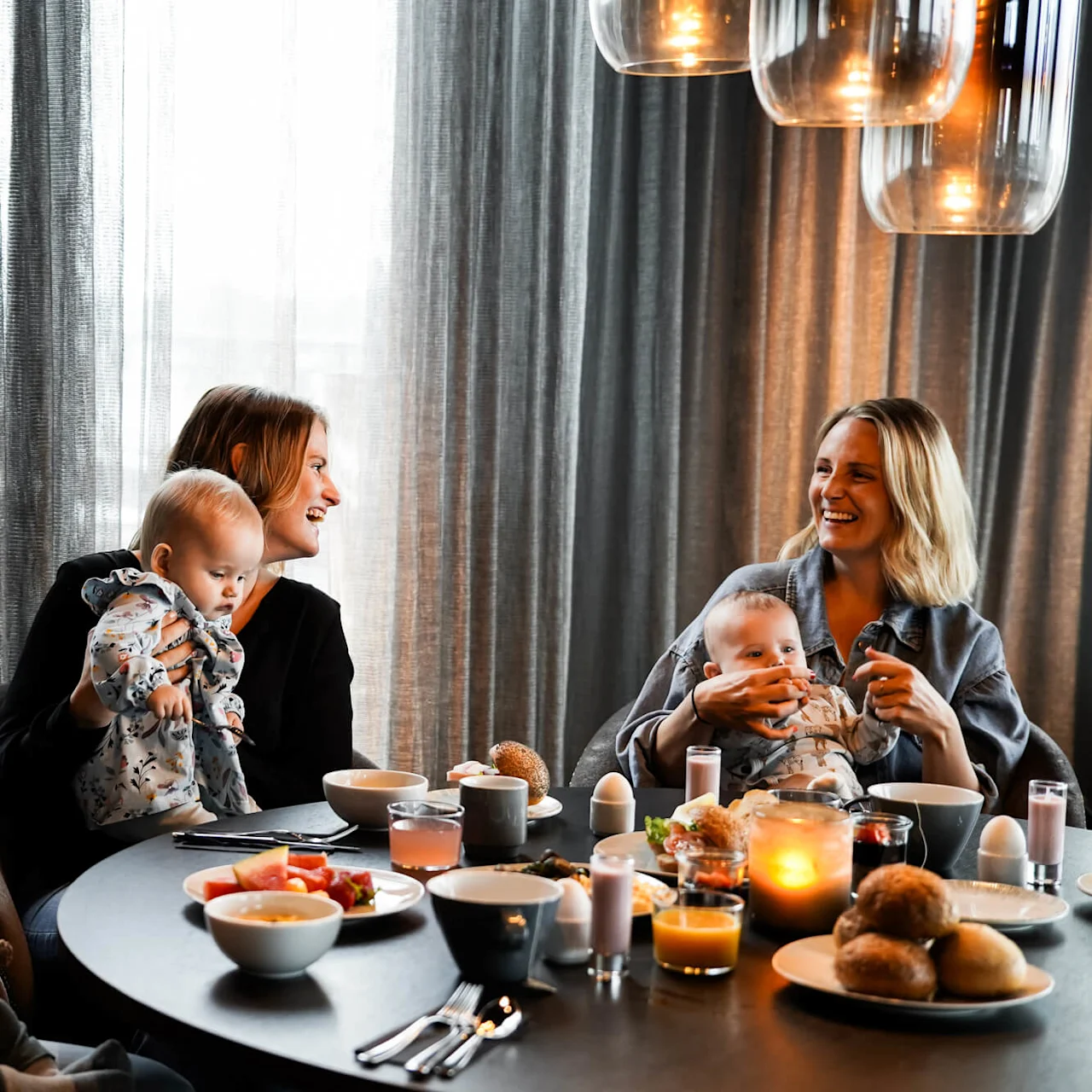 Tre kvinder sidder ved et rundt bord, hver holder en baby. De smiler og taler sammen. Bordet er dækket med morgenmad som juice, brød og æg; blød belysning hænger over.