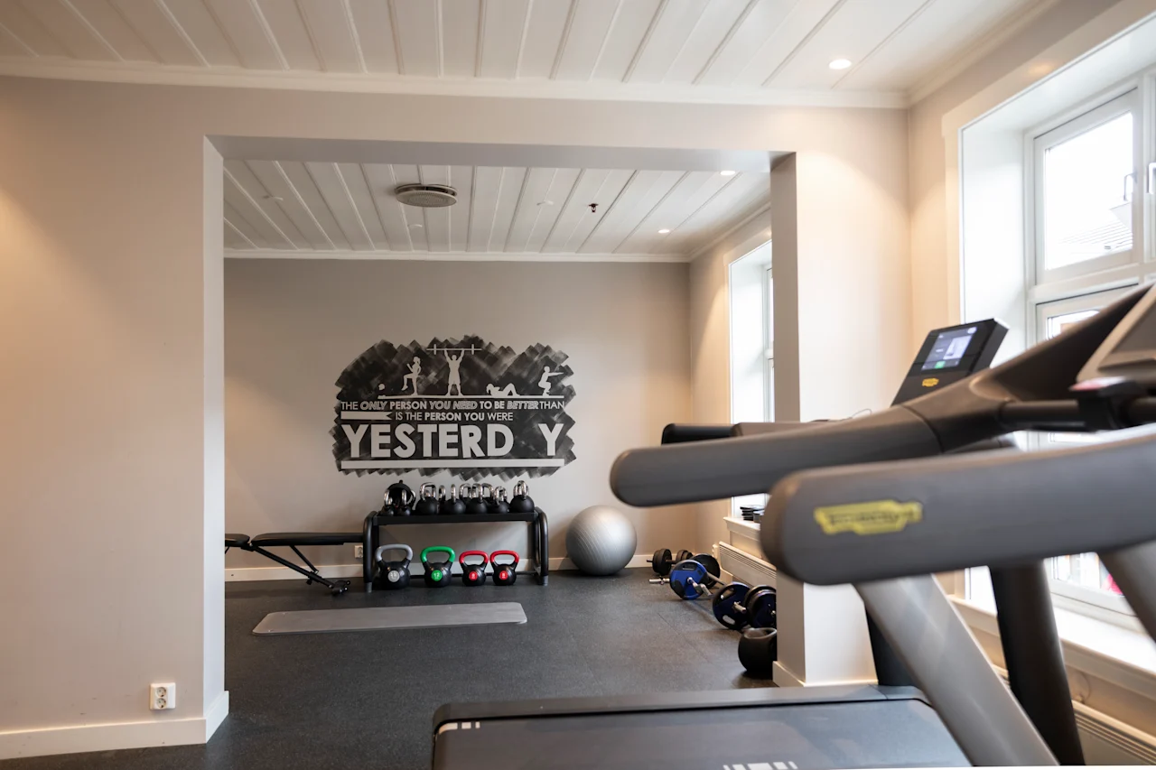 Et løbebånd står foran flere fitnessudstyr som kettlebells og en balancebold i et lyst træningsrum. På væggen står der: "THE ONLY PERSON YOU NEED TO BE BETTER THAN IS THE PERSON YOU WERE YESTERDAY."