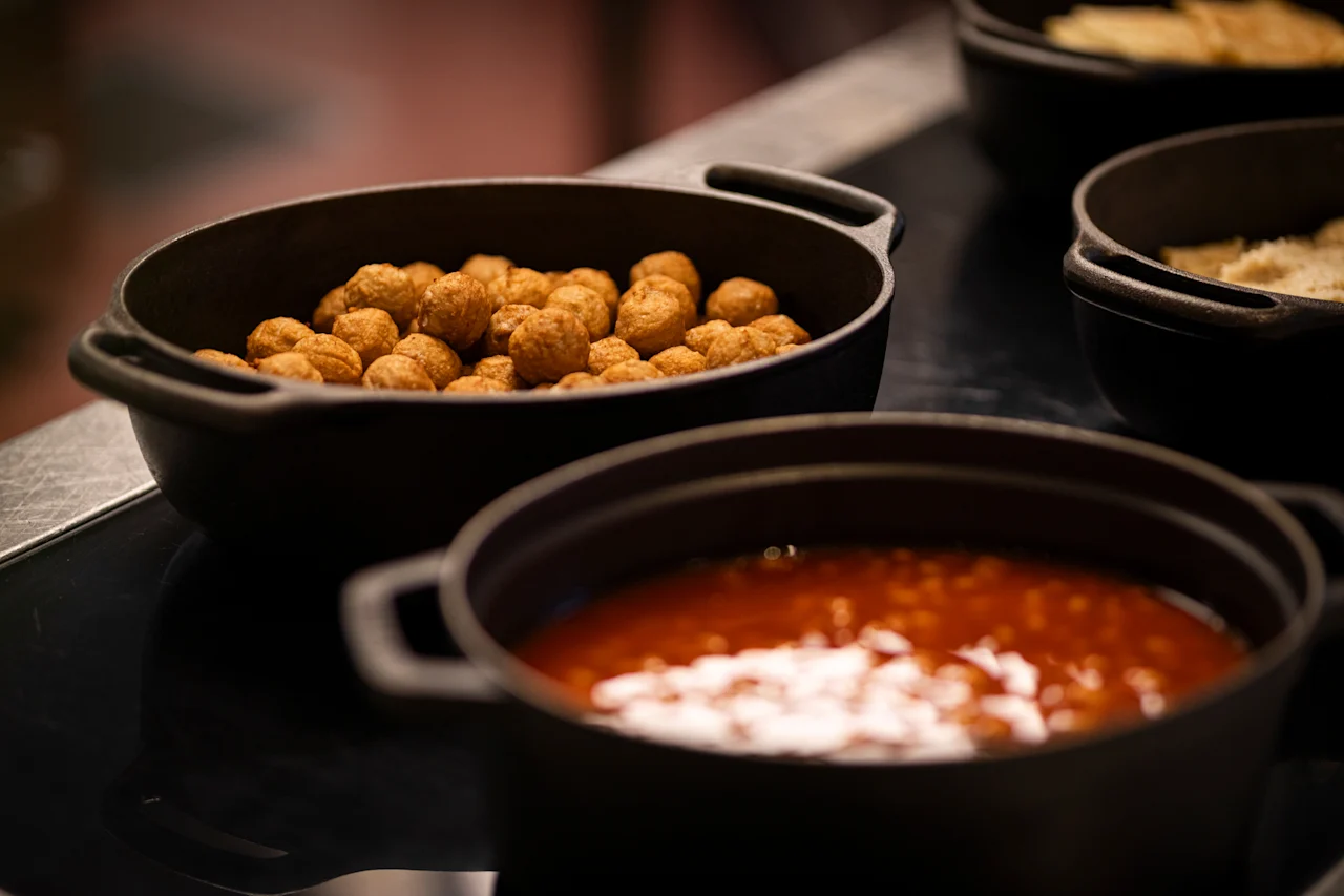 A close-up of a buffet with black pots filled with hot food, including crispy fried items and a rich red stew.