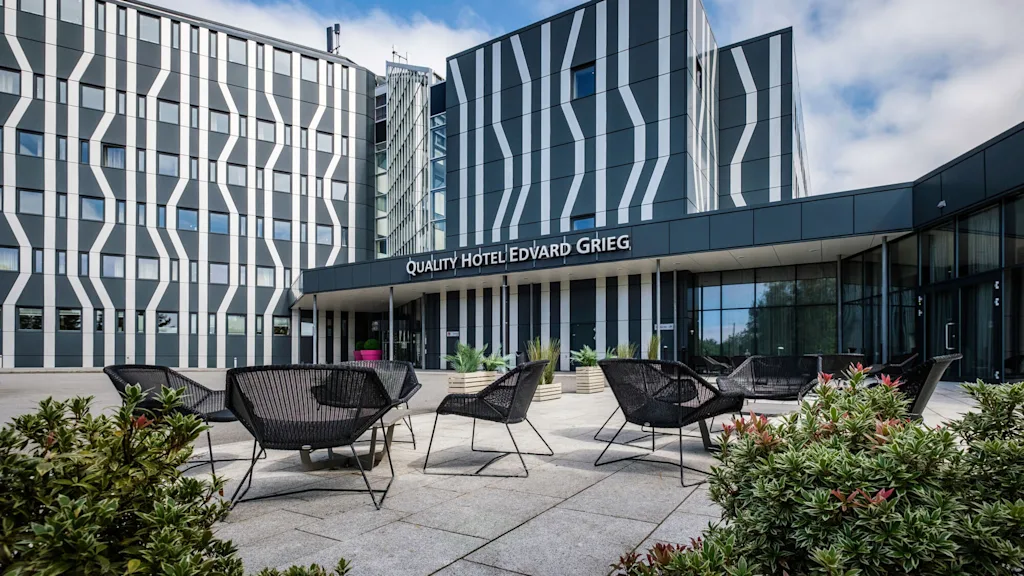 Modern hotel building with striped facade, displaying sign "QUALITY HOTEL EDVARD GRIEG", surrounded by outdoor seating and green plants under a partly cloudy sky.