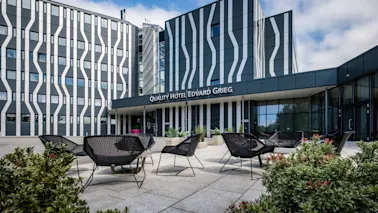 Moderne hotellbygning med stripet fasade, som viser skiltet "QUALITY HOTEL EDVARD GRIEG", omgitt av utemøbler og grønne planter under en delvis skyet himmel.