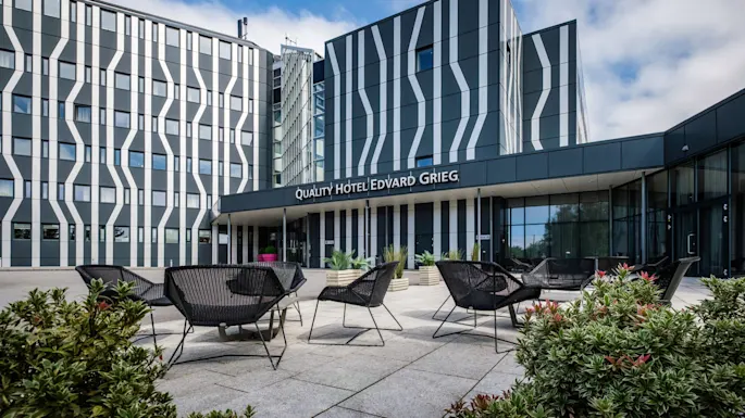 Modern hotel building with striped facade, displaying sign "QUALITY HOTEL EDVARD GRIEG", surrounded by outdoor seating and green plants under a partly cloudy sky.