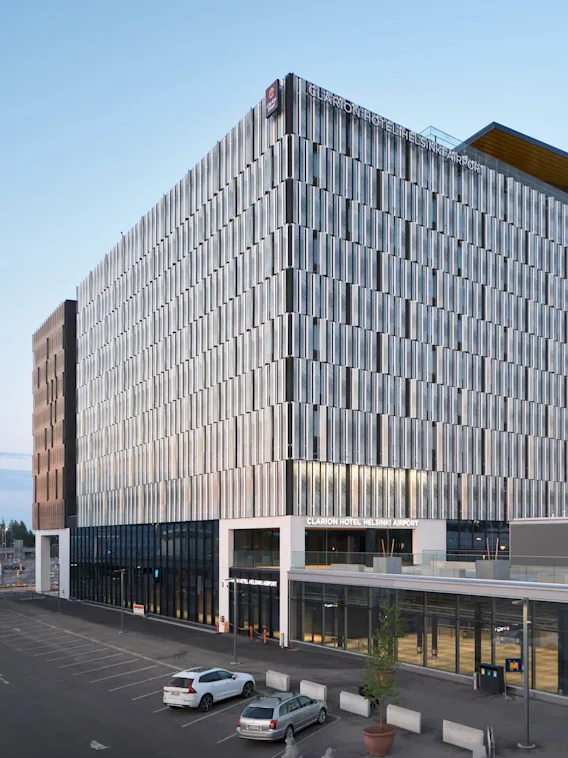 Nykyaikainen, lasiseinäinen hotellirakennus kyltillä "Clarion Hotel Helsinki Airport"; vieressä rautatieasema. Kaksi pysäköityä autoa ja kirkas sininen taivas luovat rauhallisen kaupunkinäkymän.