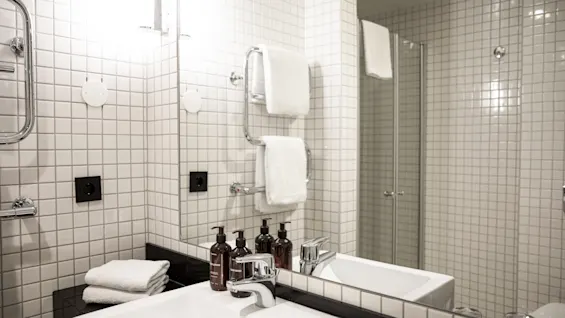 Tvättställ med tre bruna flaskor och vikta handdukar, omgivet av vita kakelväggar. En stor spegel reflekterar en handdukshållare med ytterligare handdukar, vilket förstärker den moderna badrumskänslan.