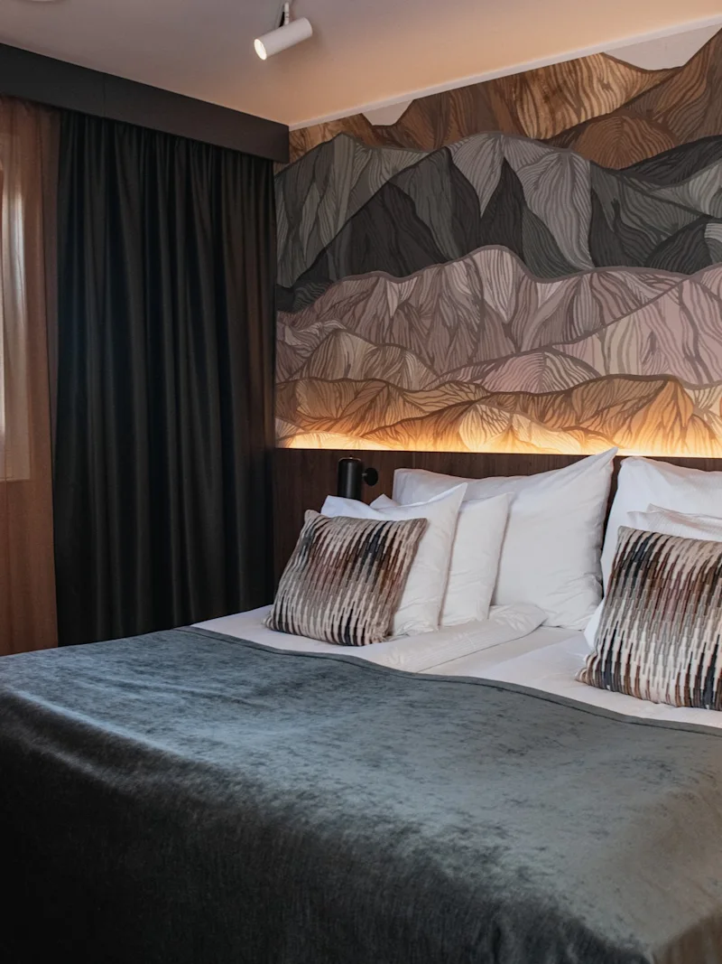 Ett mysigt hotellrum med en stor säng med grå filt, en bergsinspirerad väggmålning och ett fönster med gardiner.
