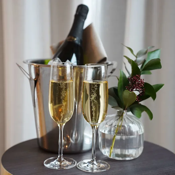 Champagne i flöjter med en flaska i en ishink och en vas med grönt, på ett träbord framför draperade gardiner.