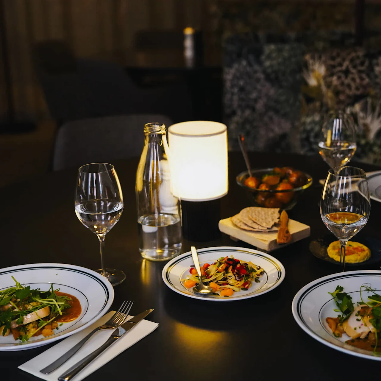 En svagt upplyst matbord med tallrikar med gourmetmat, inklusive grönsaker och färgglada garneringar, omgivna av vinglas, vattenflaska, liten lampa och olika snacks.