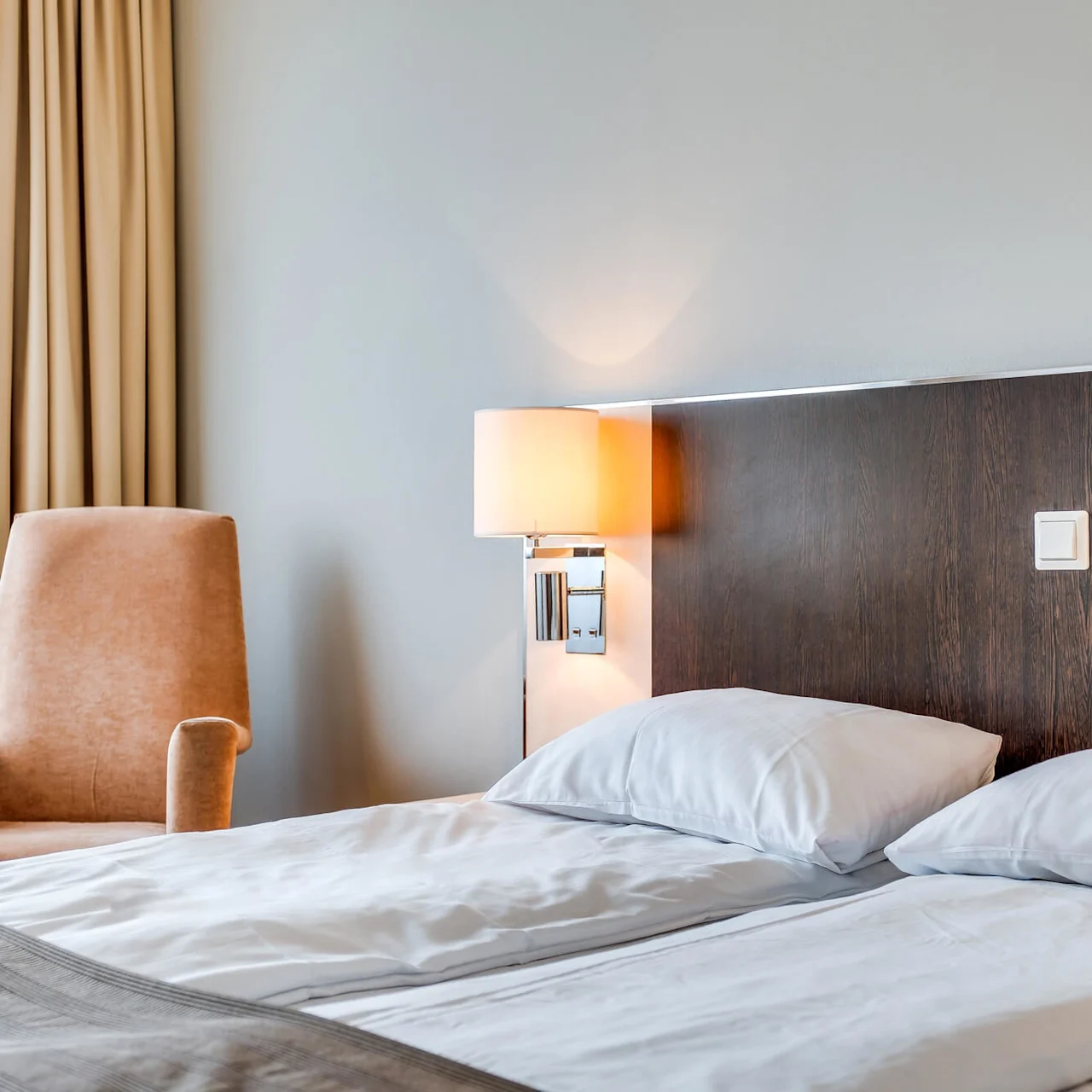A neatly made bed with a tan armchair beside it, illuminated by a lamp, in a calm hotel room.