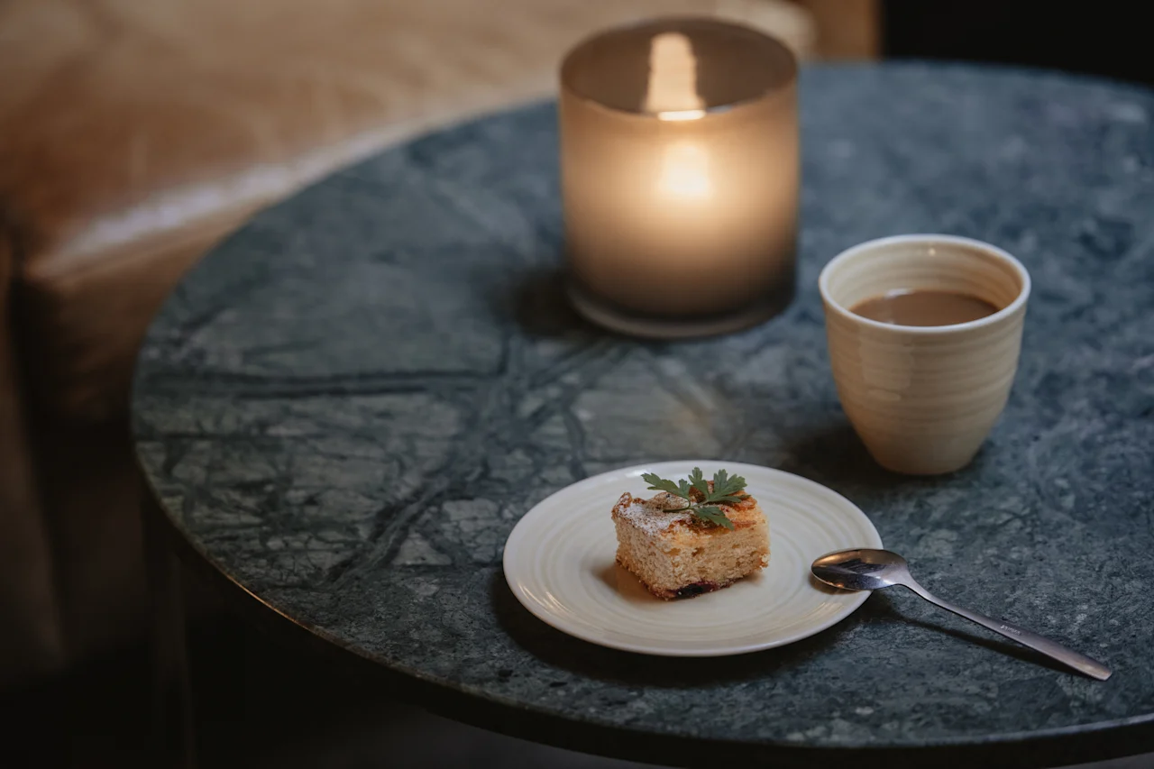 En koselig scene med et kakestykke, en kopp kaffe og et tent lys på et marmorbord, som antyder en avslappende pause.