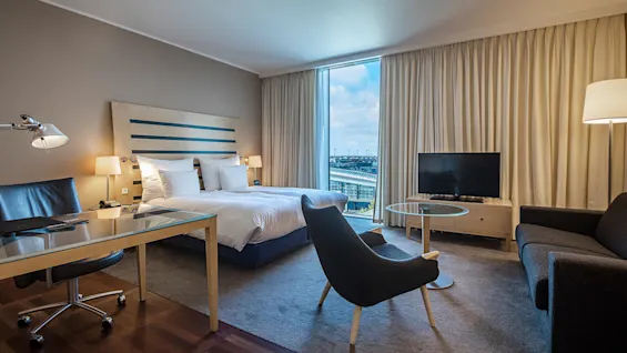 Ett hotellrum har en säng, skrivbord med stol och soffa runt en TV. Stort fönster med gardiner avslöjar en stadsvy, vilket förstärker den moderna inredningen.