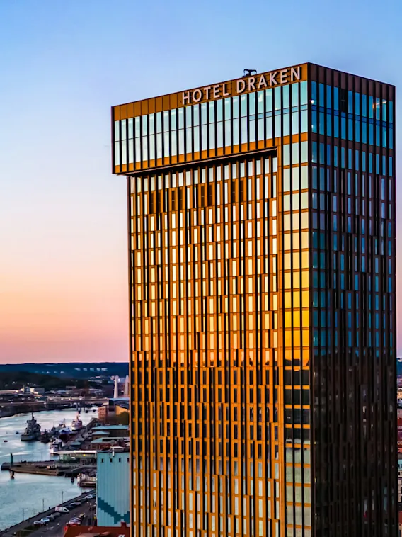 A tall glass building labeled "Hotel Draken" reflects a sunset sky over a waterfront cityscape. Nearby, a river flows alongside urban buildings and docks.