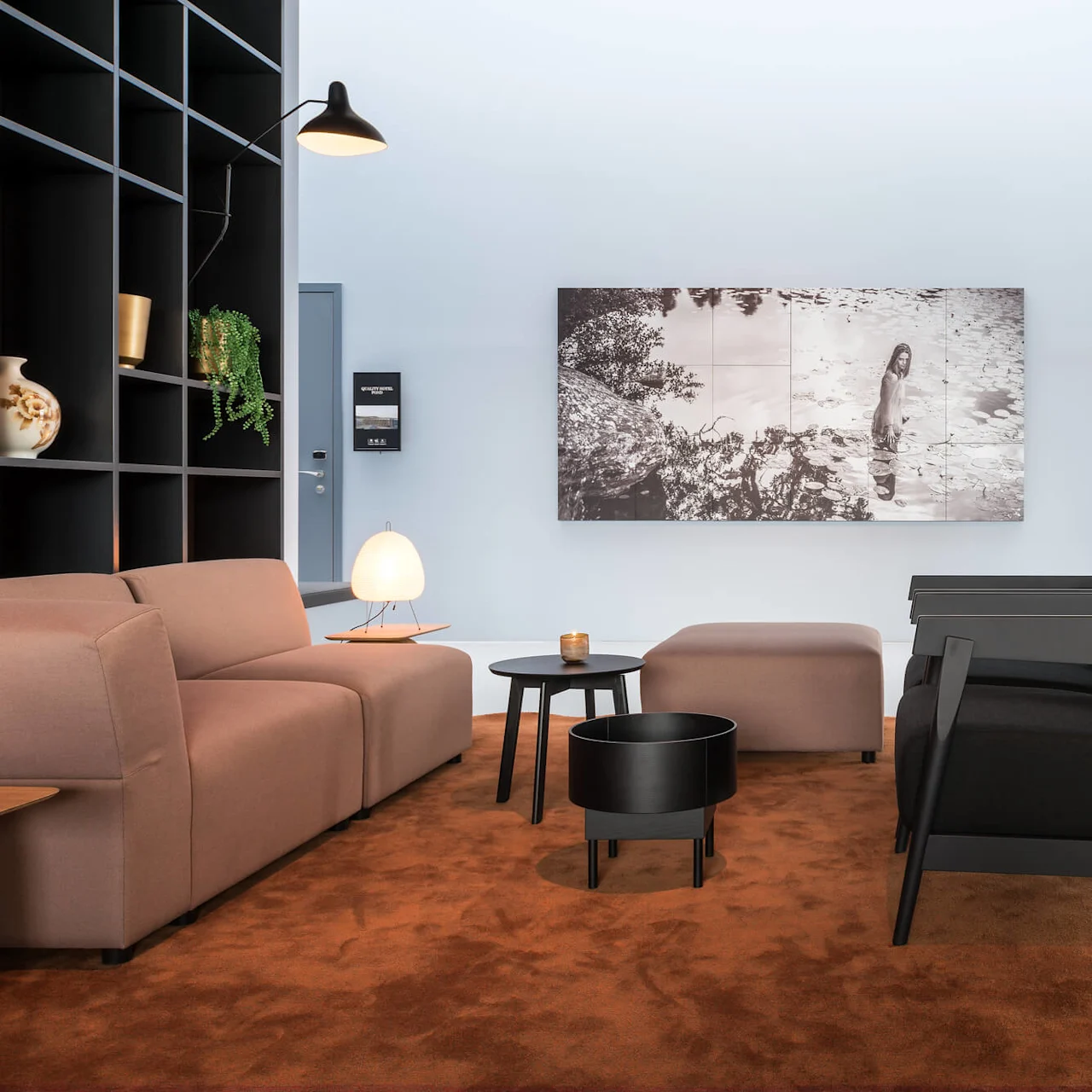 A modern lounge area features beige and black sofas arranged on a brown carpet, with small tables and decor items. On the wall, a large monochrome photograph depicts a natural landscape.