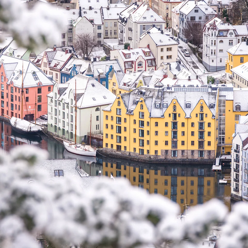 Färgglada, snöklädda byggnader kantar en lugn vattenfront och reflekteras i vattnet. Snötäckta grenar inramar förgrunden, vilket ger djup åt den vintriga stadsbilden.