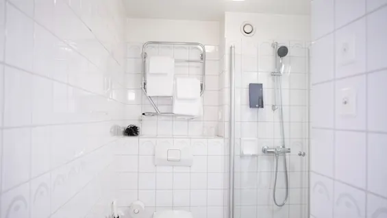 Toilet og bruser i et hvidflisebelagt badeværelse. Håndklæder hænger på et stativ over toilettet. En lampe i loftet og en sæbedispenser ved bruseren.