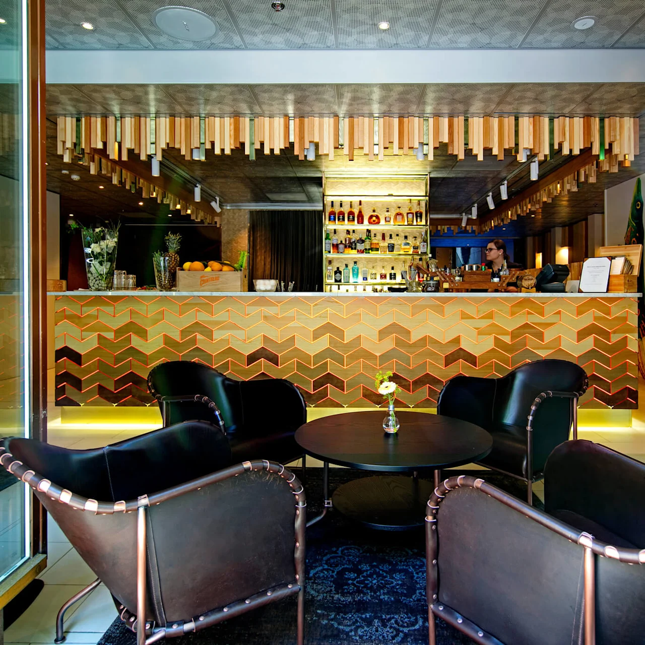 Tyylikäs baari värikkäällä siksak-kuviollisella tiskillä, tuoleja ja pöytiä etualalla. Baarimikko työskentelee tiskin takana, pullot näkyvillä. "KL7 Bar & Lounge" näkyy lasissa.
