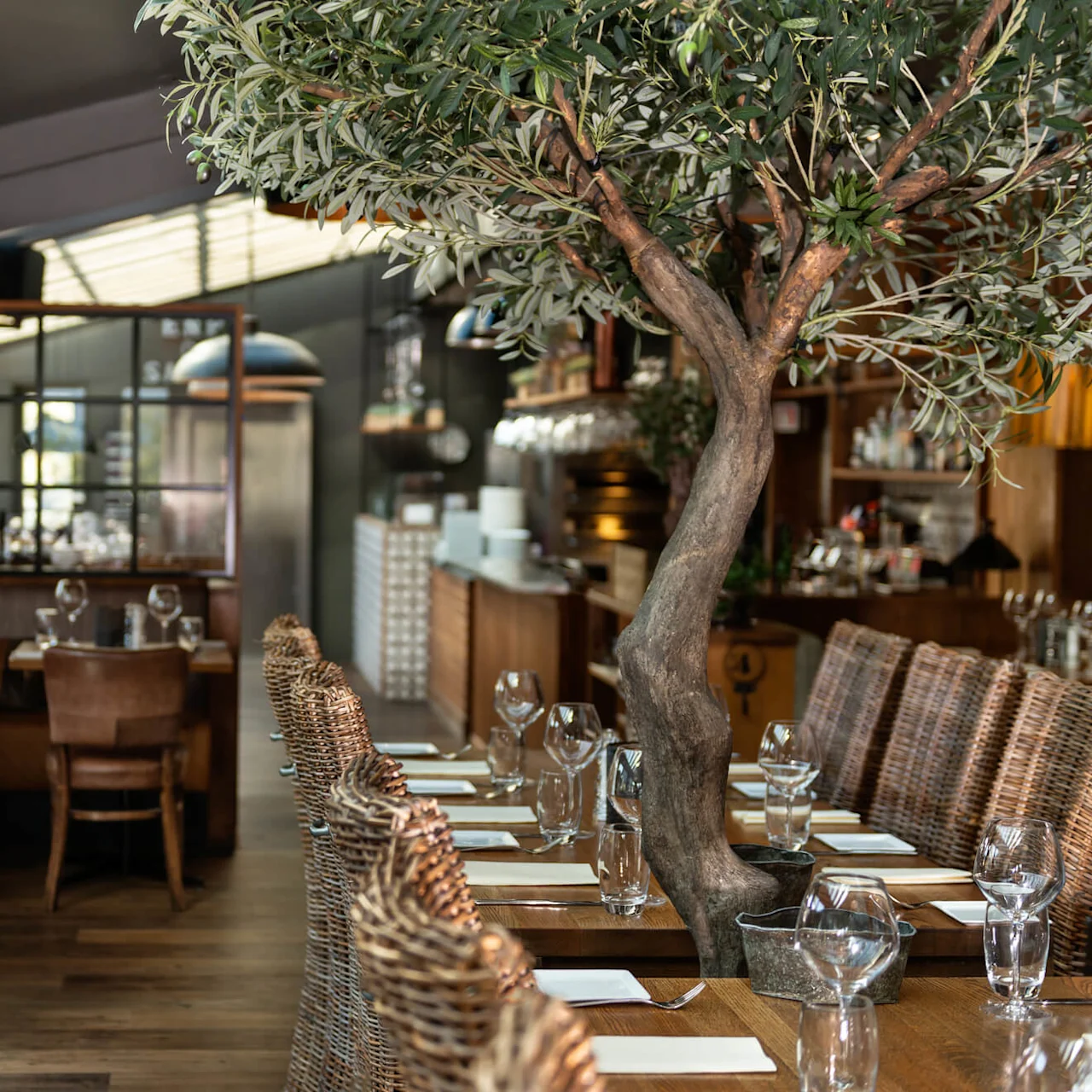 Ett stort krukträd står som mittpunkt bland dukade bord med glas och rottingstolar i en varmt belyst, elegant restaurang med trägolv och omgivande belysning.