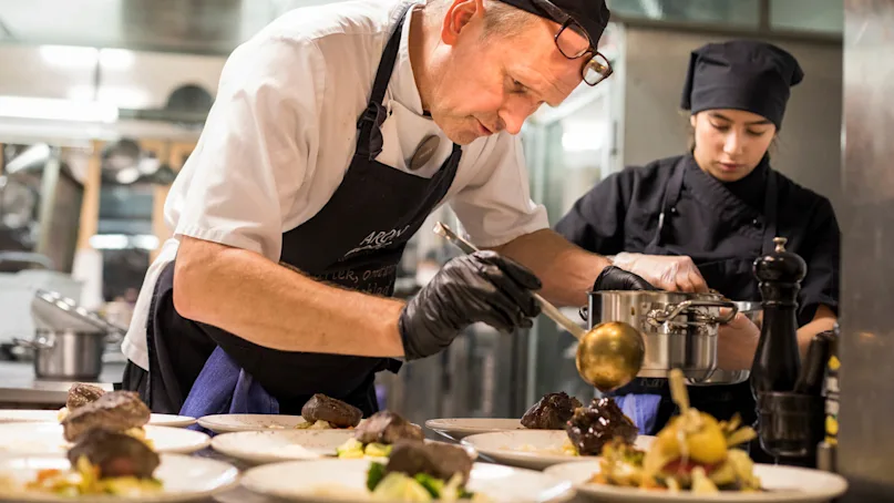 A chef garnishes several dishes in a busy kitchen, with an assistant nearby preparing a sauce. The atmosphere is professional and focused.