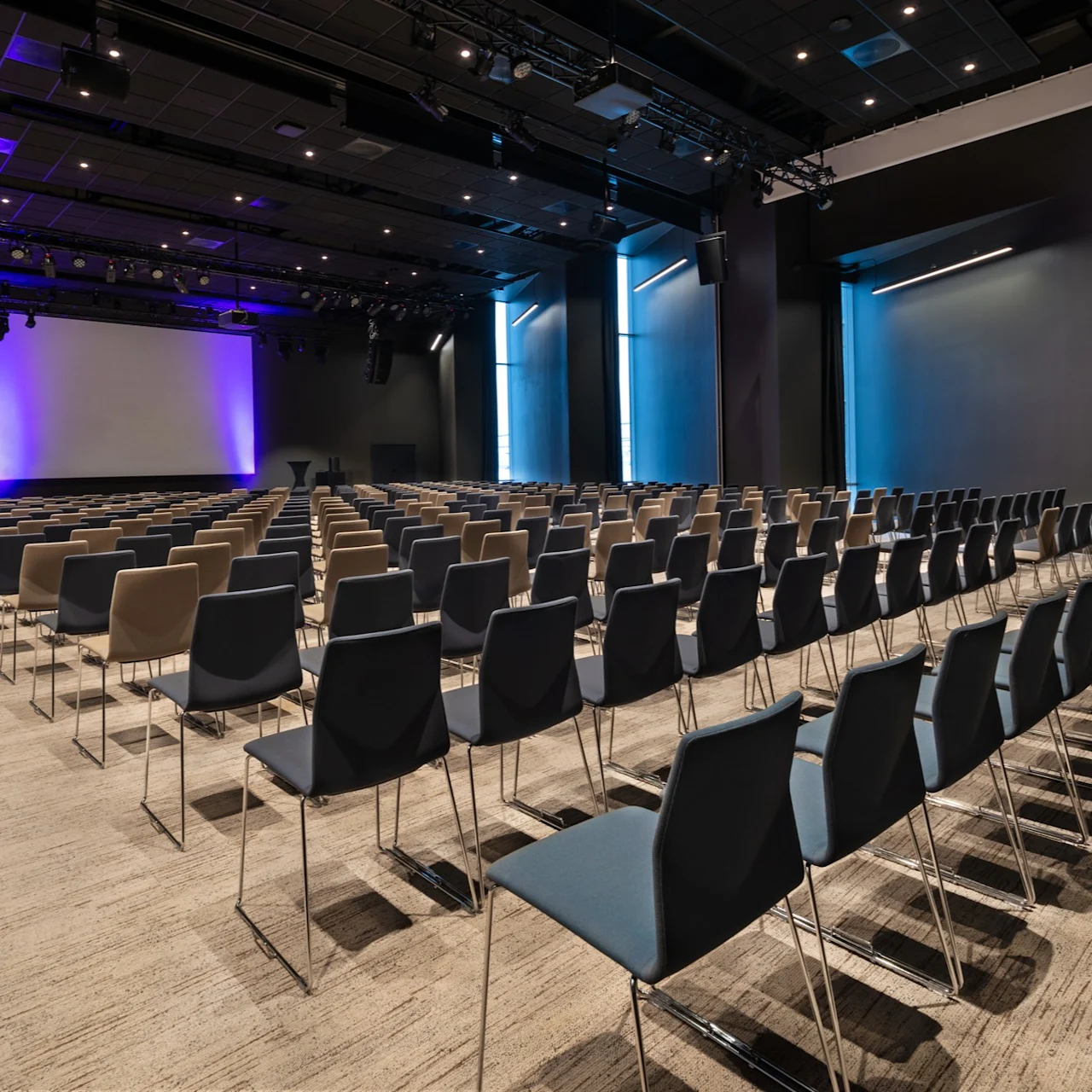 Rader av tomme, svarte stoler vender mot en stor skjerm med blå belysning i et romslig, moderne konferanserom.