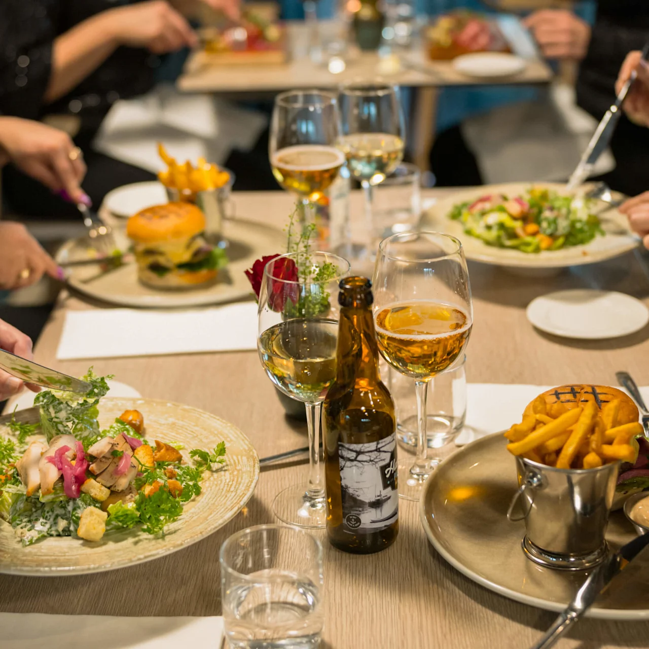 Middagsfat med sallader och hamburgare avnjuts av personer runt ett träbord, tillsammans med vinglas och ölflaskor i en restaurangmiljö.