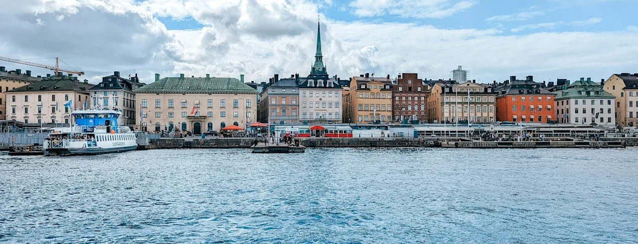 En livlig byvannfront med fargerike bygninger, et fremtredende kirketårn og en ferge på vannet under en skyet himmel.