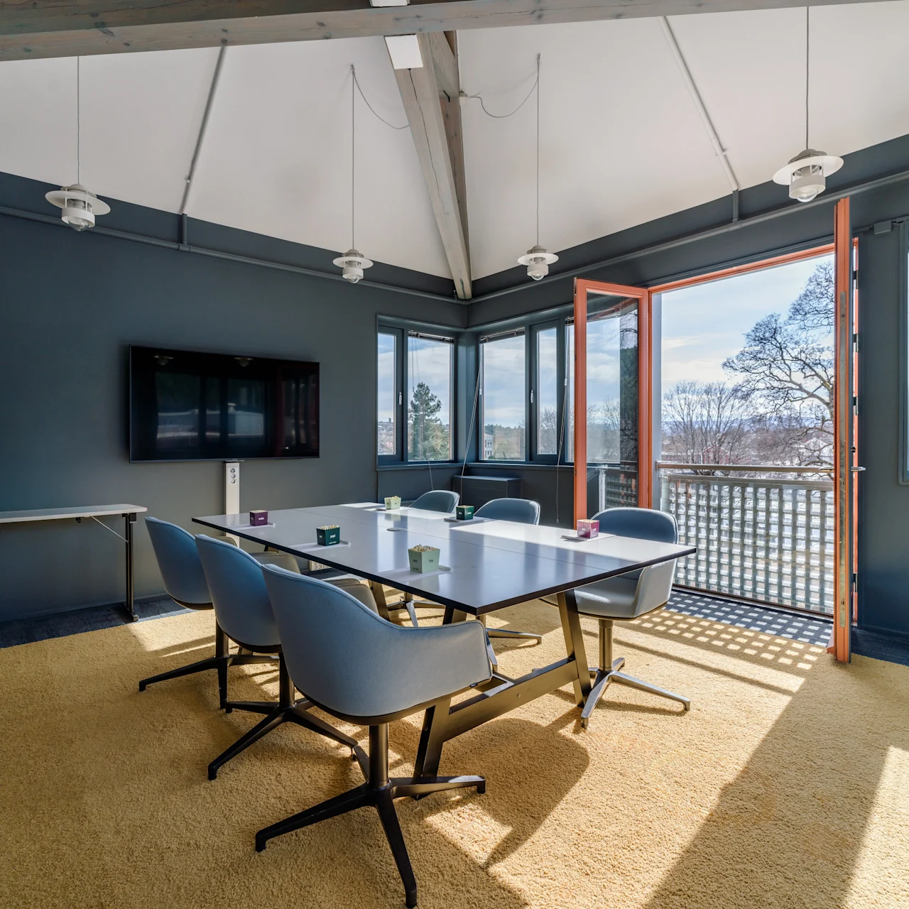 A conference table with six blue chairs is situated in a bright room. Large windows and an open door reveal a sunny view of trees outside, and light fixtures hang overhead.