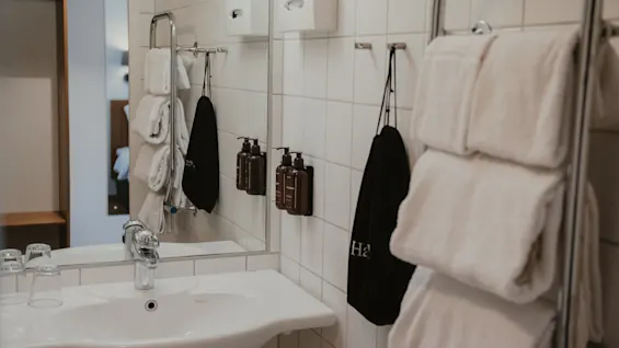 Siisti ja moderni hotellin kylpyhuone, jossa on pesuallas, peili, puhtaita valkoisia pyyhkeitä ja hygieniatuotteita. Musta pyyhe, jossa 'Ha', näkyy.