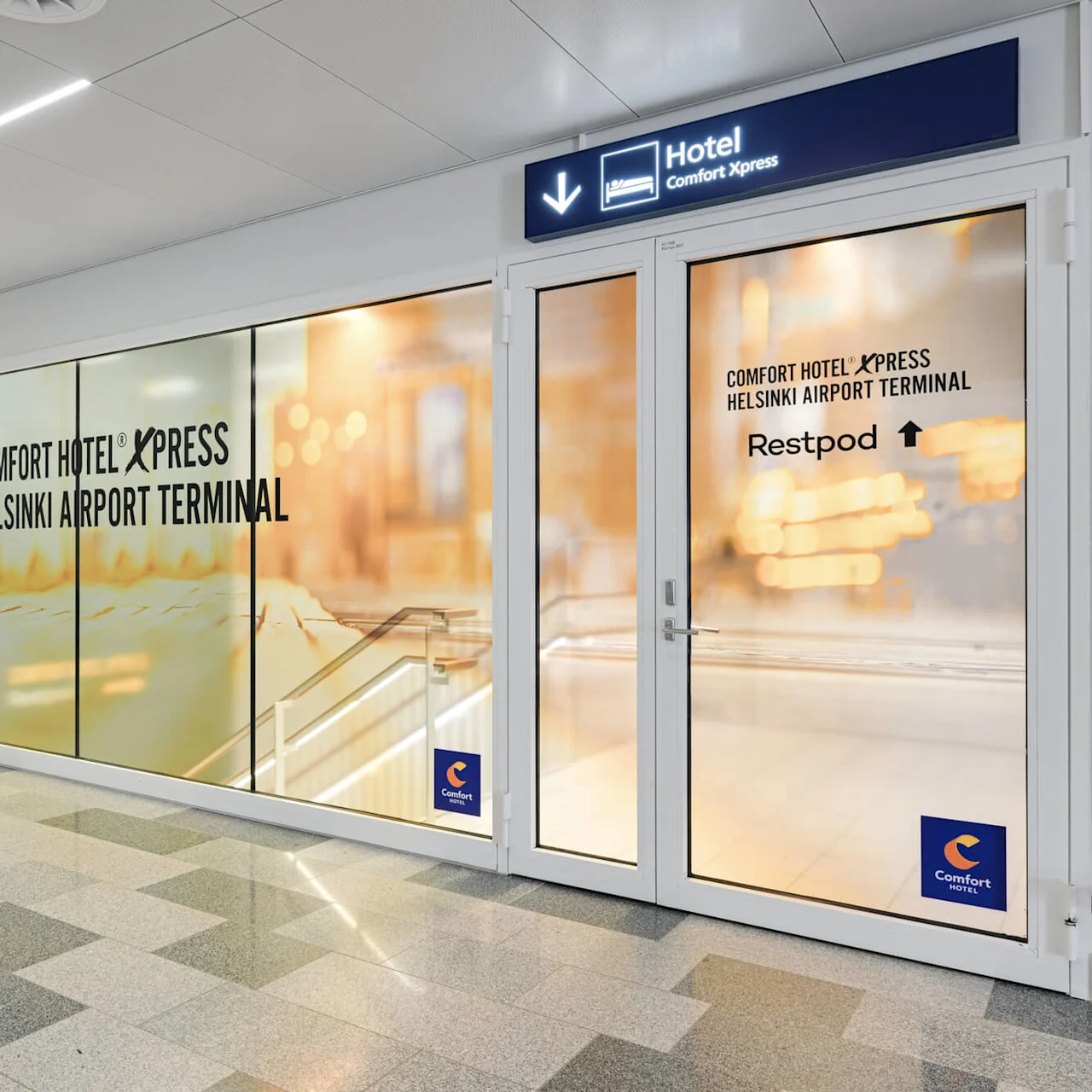 Comfort Hotel Xpress-entré med stora fönster och dörrar, visar texten: "COMFORT HOTEL XPRESS HELSINKI AIRPORT TERMINAL" och "Restpod." Ligger i en ljus, kaklad flygplatskorridor.