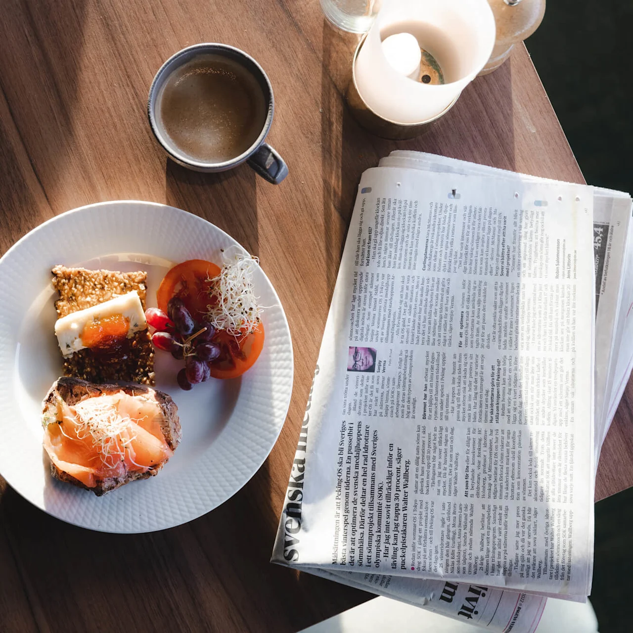 En vit tallrik med kex och ost, serverad med vindruvor och tomat. I närheten en kaffekopp och en öppnad tidning med titeln "Svenska".
